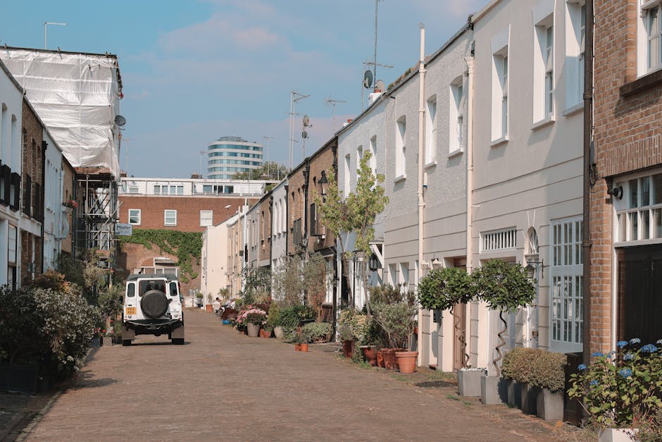 A narrow cobblestone street lined with a mix of Victorian terraced houses featuring white and brick facades, with small front gardens containing neatly trimmed shrubs, potted plants, and flowering bus