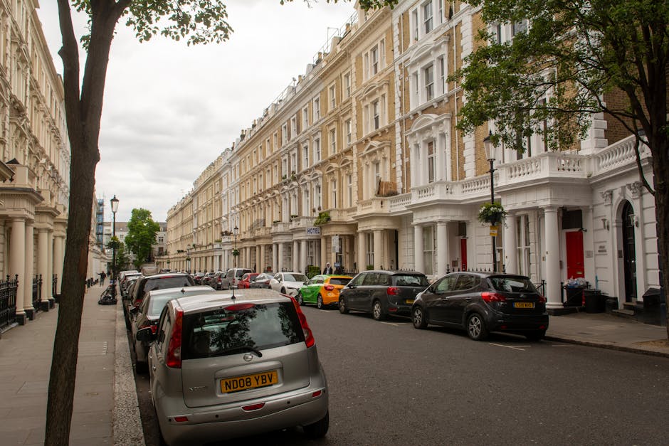 A narrow cobblestone street lined with a mix of Victorian terraced houses featuring white and brick facades, with small front gardens containing neatly trimmed shrubs, potted plants, and flowering bus
