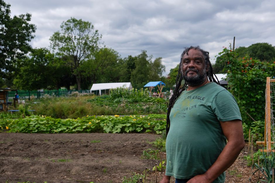 Two individuals are engaged in garden maintenance within a shared green space at Weavers Fields, with a white picket fence and modern apartment buildings visible in the background. They are positioned